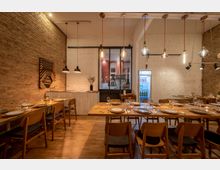 Innenraum eines gemütlichen Restaurants mit Holzböden und Tischen, die mit Geschirr und Gläsern gedeckt sind. Die Wände bestehen aus freiliegendem Ziegelstein und weiß gestrichenem Mauerwerk, mit hängenden Lampen und einem beleuchteten Getränkekühlschrank im Hintergrund.