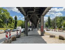 Blick unter eine Hochbahnbrücke aus Stahl mit Gehweg aus Kopfsteinpflaster, flankiert von einer Straße mit Autos und Bauabsperrungen. Im Hintergrund sind Bäume, Wohnhäuser und ein blauer Himmel mit Wolken zu sehen.