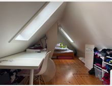 Dachzimmer mit schrägen weißen Wänden und zwei großen Dachfenstern, Holzfußboden und einem kleinen Bett am hinteren Ende. Links steht ein weißer Schreibtisch mit Stuhl, rechts Regale und ein Schrank mit Kleidung und Aufbewahrungsboxen.