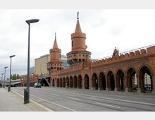 Rote Backsteinbrücke mit zwei Türmen im neugotischen Stil, bekannt als die Oberbaumbrücke in Berlin, mit umliegenden modernen Gebäuden im Hintergrund und einer freien Straße im Vordergrund. Die Arkaden der Brücke zeigen gotische Spitzbögen, und die Türme haben konische Dächer.