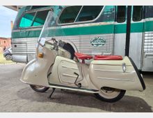 Ein cremefarbener Motorroller mit hoher Windschutzscheibe und rotem Sitz steht auf einem Betonboden unter einem überdachten Bereich. Im Hintergrund ist die Seite eines grün-silbernen, genieteten Oldtimerbusses mit großen Fenstern zu sehen, daneben ein geparktes Auto und Backsteingebäude.