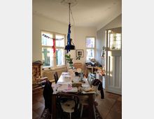 Dining room with a large wooden table covered in papers and household items, surrounded by mismatched chairs. The space features a ceiling light with colorful streamers, stained glass on a door, and "Happy Birthday" decorations on the windows.