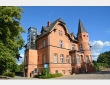 Backsteinbau im neugotischen Stil mit turmartigen Elementen und reich verzierten Giebeln, ergänzt durch einen modernen Glasaufzug an der linken Seite. Das Gebäude steht in einer grünen Umgebung mit Kopfsteinpflaster im Vordergrund und klarem Himmel im Hintergrund.