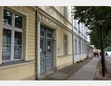 Straßenansicht mit einem beige-gelben historischen Gebäude mit verzierten Türen und Fensterrahmen. Rechts führt ein gepflasterter Bürgersteig entlang einer Baumreihe, im Hintergrund sind Fußgänger und ein Fluss erkennbar.