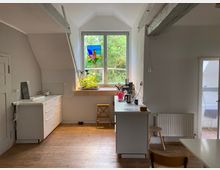 Helles Zimmer mit Holzfußboden und einer kleinen Küche auf der rechten Seite; ein großes Fenster mit bunter Glasdekoration und Blick ins Grüne befindet sich an der Stirnwand. Sichtbare Holzbalken an der weißen Decke und eine Tür im Hintergrund führen zu einem weiteren Raum.