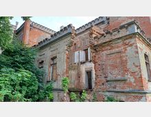 Verfallenes Backsteingebäude mit teilweise eingestürzten Wänden und beschädigten Fenstern, umgeben von dichtem Grün. Die Fassade zeigt Spuren von Putzabplatzungen und eine Zinnenstruktur an der oberen Kante.