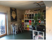 Ein Raum mit Betonwänden und grünem Boden, ausgestattet mit einem Regal, das zahlreiche Farbbehälter und Arbeitsmaterialien enthält. Links ist ein großes Fenster mit Holzrahmen, und daneben stehen Klappleitern sowie eine großformatige Illustration an der Wand; im Vordergrund befinden sich ein Drucker und ein großer Zimmerpflanzentopf.