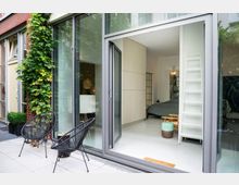 Moderne Wohnung mit einer großen Glasfront und Schiebetür, die einen Blick in ein helles Schlafzimmer mit minimalistischer Einrichtung bietet. Außen befinden sich eine gepflasterte Terrasse, zwei schwarze Stühle und eine mit Pflanzen bewachsene Ziegelwand.