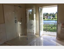 Ein luxuriöses Badezimmer mit einer verglasten Dusche und glänzenden Marmorfliesen; durch eine geöffnete Doppeltür mit zierlichem Balkongeländer ist ein Garten mit Fluss und Häusern im Hintergrund sichtbar.