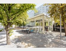 Ein modernes Cafégebäude mit großen Glaswänden und flachem Dach, umgeben von Bäumen auf einem gepflasterten Platz. Draußen stehen Tische mit Sonnenschirmen, und im Hintergrund ist ein mehrstöckiges Wohngebäude zu sehen.