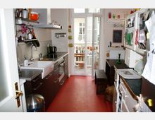 Schmale Küche mit rotem Boden, dunklen Unterschränken, Edelstahl-Arbeitsflächen und einer weißen Spüle. Am Ende des Raumes führen Glastüren auf einen kleinen Balkon mit Tisch und Stühlen.