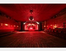 Ein Innenraum eines Veranstaltungsraums mit dunklem Holzboden, roter Beleuchtung und einer Bühne im Hintergrund. An den Seiten befinden sich gepolsterte Sitzbänke und eine Discokugel hängt an der Decke.