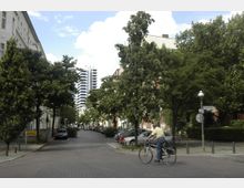 Straßenszene in einer Wohngegend mit gepflasterter Fahrbahn, Reihen von parkenden Autos und Bäumen, die den Straßenrand säumen. Im Hintergrund ist ein modernes Hochhaus zu sehen, und im Vordergrund fährt ein Mann auf einem Fahrrad vorbei.