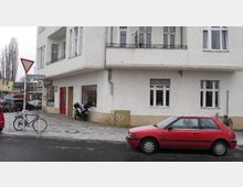Ecke eines Wohngebäudes mit weißer Fassade und mehreren Fenstern, darunter ein Geschäft mit Schaufenster im Erdgeschoss. Davor stehen ein rotes Auto, Fahrräder und ein Vorfahrt-achten-Schild an der Straße.
