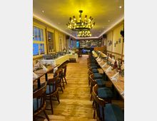 Ein festlich dekorierter Gastraum mit Holzboden, langen gedeckten Tischen und gepolsterten Holzstühlen. Im Hintergrund befindet sich eine beleuchtete Theke mit Flaschenregalen, an der Decke hängt ein großer, moderner Kronleuchter mit warmem Licht.