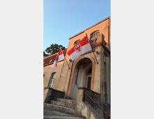 Ein zweistöckiges, beiges Gebäude mit einem Eingang in Rundbogenarchitektur und einer Steintreppe. Zwei Flaggen mit dem Wappen des Bundeslandes Brandenburg hängen an Fahnenstangen links und rechts des Eingangs.