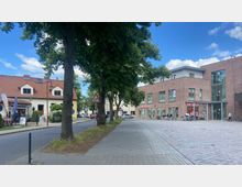 Straßenszene mit Bäumen und Gehwegen; links ein Gebäude mit rotem Ziegeldach, das wie ein Café oder Geschäft aussieht, rechts ein moderner Gebäudekomplex aus rotbraunem Backstein mit einem "Rossmann"-Ladeneingang. Autos und Fahrräder sind vor den Gebäuden geparkt, der Himmel ist teils bewölkt.