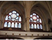 Innenraum einer Kirche mit gotischer Architektur, dekoriert mit großen, farbigen Rosettenfenstern und Spitzbogenfenstern. Die hohen Säulen und Gewölbedecken tragen die Struktur, und eine umlaufende Empore mit verzierten Balustraden ist sichtbar.