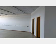 Leerer Innenraum mit weiß gestrichenen Wänden, einer abgehängten Decke mit Leuchtstofflampen und zwei Türen mit gelben Rahmen. Der Boden ist glatt und grau, und an der Wand verlaufen Steckdosenleisten.