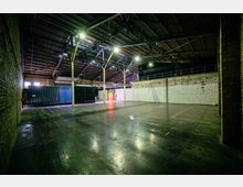 Geräumige Industriehalle mit hohen Decken aus Metall, freiliegenden Ziegel- und Betonwänden sowie einem großen dunklen Container an einer Seite. Das reflektierende Betonboden, eine Metallbühnenkonstruktion an der Decke und spärliche Beleuchtung definieren die leere Innenfläche.