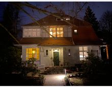 Zweistöckiges Wohnhaus mit weißer Fassade und rotem Satteldach, beleuchtet in der Dämmerung. Die Fenster sind teilweise erleuchtet, umliegende Bäume und Büsche rahmen das Gebäude ein.