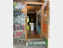 Eingangstür mit Holzrahmen und großen Glasflächen, die dicht mit Graffiti und Tags besprüht sind. Durch die offene Tür ist ein schmaler Innenraum mit Holzboden, dunklen Wänden und einem hellen Tresen sowie Lampen sichtbar.