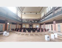 Ein großes, altes Hallenbad mit hohen Decken, Oberlichtern und gelben Wänden, umgewandelt in einen Veranstaltungsraum mit aufgestellten Stühlen in Reihen. Entlang der Wände befinden sich ehemalige Umkleidekabinen mit hellblauen Türen; oben führt eine umlaufende Galerie mit Geländern entlang der Wände.