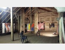 Dachboden einer alten Scheune oder Lagerhalle mit einer Kunstinstallation. Abstrakte Gemälde sind zwischen Holzbalken aufgehängt, und Plastikstühle sind auf dem Holzboden verteilt.