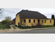 Gelbes Einfamilienhaus mit Satteldach an einer asphaltierten Straße, umgeben von Büschen und kleinen Bäumen. Auf der linken Seite führt ein unbefestigter Weg durch ein Metalltor zu einem roten Backsteingebäude im Hintergrund.