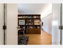 Ein modernes Arbeitszimmer mit einem offenen Regal aus dunklem Holz, das Bücher und Dekorationsstücke enthält. Der Raum hat helle Wände, einen Holzboden, einen schwarzen Bürostuhl vor einem Schreibtisch und eine Schiebetür auf der rechten Seite.