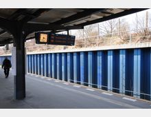 Eine überdachte U-Bahn-Station mit Gleis, blauer Metallwand im Hintergrund und einer Anzeigetafel, die Abfahrtszeiten für die Linie U3 anzeigt. Im Freien sind kahle Bäume und eine niedrige Metallbegrenzung sichtbar.
