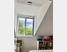 Zimmer mit Dachschräge, weiß gestrichenen Wänden und einem offenen Fenster mit Blick auf grüne Bäume und bewölkten Himmel. Im Raum befinden sich ein Bücherregal mit Büchern und Ordnern sowie gerahmte Bilder, die an der Wand lehnen.