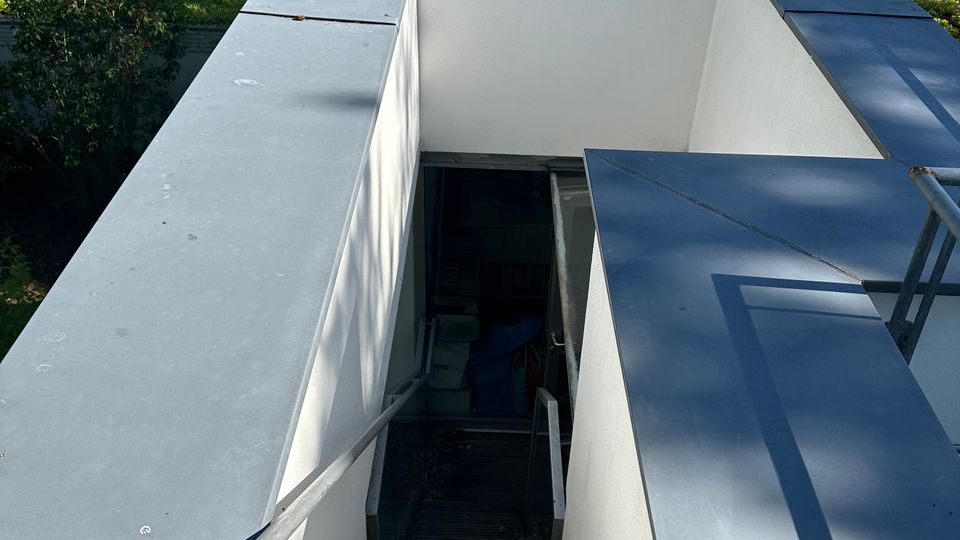 Blick auf eine schmale, außenliegende Treppe, die zwischen weiß verputzten Mauern mit grauen Metallabdeckungen nach unten führt. Oben verläuft ein Geländer entlang einer begrünten Dachfläche; im Hintergrund stehen ein Haus mit Ziegeldach und umgebende Bäume.
