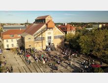 Luftaufnahme eines historischen Gebäudekomplexes mit Backsteinfassade und Satteldach, umgeben von Bäumen und niedrigen Häusern. Vor dem Hauptgebäude befinden sich zahlreiche Menschen bei einer Veranstaltung mit Tischen und Ständen im Freien.