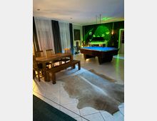 Großer Wohn- und Spielbereich mit gefliestem Boden, Esstisch aus Holz mit Bänken und Stühlen sowie einem Billardtisch. Im Hintergrund hängen bodentiefe Vorhänge; eine grün beleuchtete Wand mit Dschungelmotiv und Kamin bildet einen auffälligen Akzent.