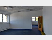 Ein leerer Büroraum mit weiß gestrichenen Wänden, blauer Teppichboden und einer Decke mit rechteckigen Paneelen sowie integrierter Beleuchtung. Links sind große Fenster mit Jalousien, rechts eine geöffnete Holztür, die zu einem angrenzenden Raum führt.