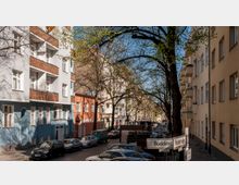 Straßenszene in einer Wohngegend mit mehrstöckigen Wohnhäusern, teils pastellfarben gestrichen, und Bäumen, deren Äste sich über die Straße spannen. Parkende Autos säumen die Kopfsteinpflasterstraße, während ein Straßenschild auf die Kreuzung von Boddinstraße und Reuterstraße hinweist.