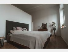 Minimalistisch eingerichtetes Schlafzimmer mit einem großen Bett mit grauem Kopfteil und mehreren Kissen. Im Hintergrund stehen eine Kommode mit Dekorationsobjekten und Pflanzen sowie ein Fenster, das natürliches Licht in den Raum lässt.