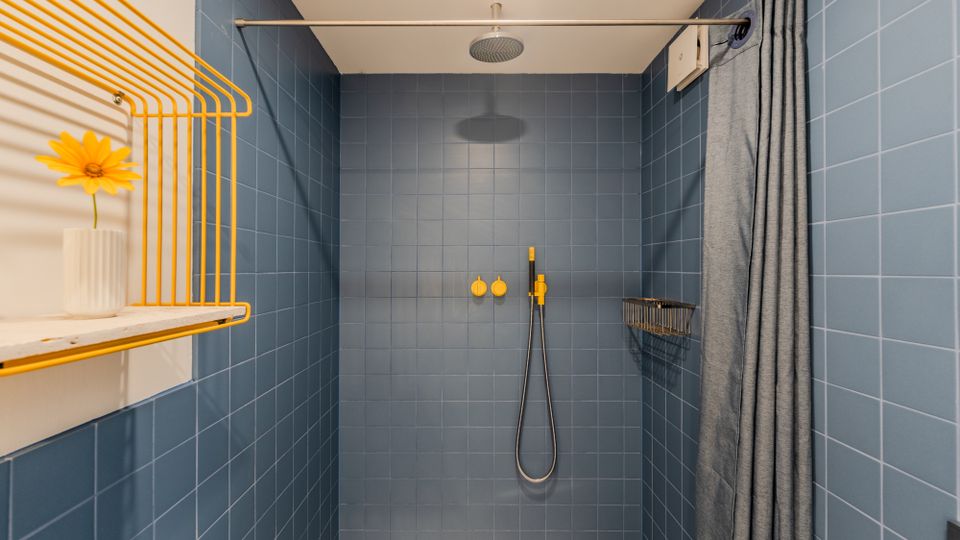 Geflieste Duschkabine mit blauen Wandfliesen, Regenduschkopf an der Decke und Handbrause mit gelben Armaturen an der Rückwand. Rechts hängt ein grauer Duschvorhang an einer Stange, links ist ein gelbes Drahtregal mit Ablage zu sehen.