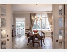 Esszimmer in einem stilvollen Altbau mit Holzboden, einem großen Holztisch und Stühlen. Ein Kronleuchter hängt zentral über dem Tisch, und große Fenster mit Vorhängen lassen Tageslicht in den Raum. Im Hintergrund ist ein weiterer Raum mit weißer Einrichtung und einem kleinen Tisch sichtbar.