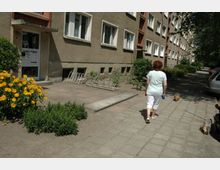Eine Wohnhausfassade mit hellbraunem Putz, mehreren Reihen von weißen Fenstern und einem Eingangsbereich mit Briefkästen. Im Vordergrund ist eine Frau mit zwei kleinen Hunden auf einem breiten Gehweg zu sehen, umgeben von Blumenbeeten und Fahrradständern.