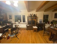 Ein Wohnraum mit Holzboden und sichtbaren Deckenbalken. Im Vordergrund ein Schlagzeug und Mikrofonstative, im Hintergrund Möbel wie ein Glasvitrinenschrank, ein Schreibtisch mit Computer und ein runder Esstisch.