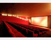 Innenansicht eines Kinosaals mit roten Polstersitzen in mehreren Reihen und einer großen, beleuchteten Leinwand vorne. Die Wände sind rot gefärbt, und dezente Lichter an den Seiten sorgen für sanfte Beleuchtung.