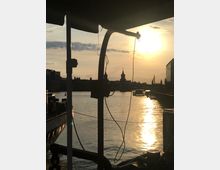 Uferansicht eines Flusses bei Sonnenuntergang, mit der Silhouette der Oberbaumbrücke im Hintergrund. Im Vordergrund sind Teile eines Bootes und lose Kabel unter einem Sonnendach zu sehen.