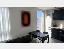 Heller Wohn- und Essbereich mit kleiner Küchenzeile, schwarzem Esstisch und Stühlen sowie dunklen Küchenschränken neben einem weißen Retro-Kühlschrank. Große Fensterfront mit Vorhängen und Glastür zum Balkon lassen viel Tageslicht herein; an der Wand hängt ein Bild.