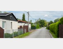 Eine schmale Wohnstraße mit einzelnen Häusern auf der linken Seite, die von niedrigen Zäunen umgeben sind. Rechts verläuft eine hohe, dichte Hecke entlang des Gehwegs, im Hintergrund sind Bäume und ein bewölkter Himmel sichtbar.