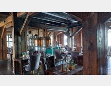 Innenraum eines rustikalen Restaurants mit sichtbaren Backsteinwänden, hölzernen Deckenbalken und Braukesseln aus Kupfer im Hintergrund. Die Einrichtung besteht aus Holztischen und gepolsterten Stühlen in einem gemütlichen Ambiente.