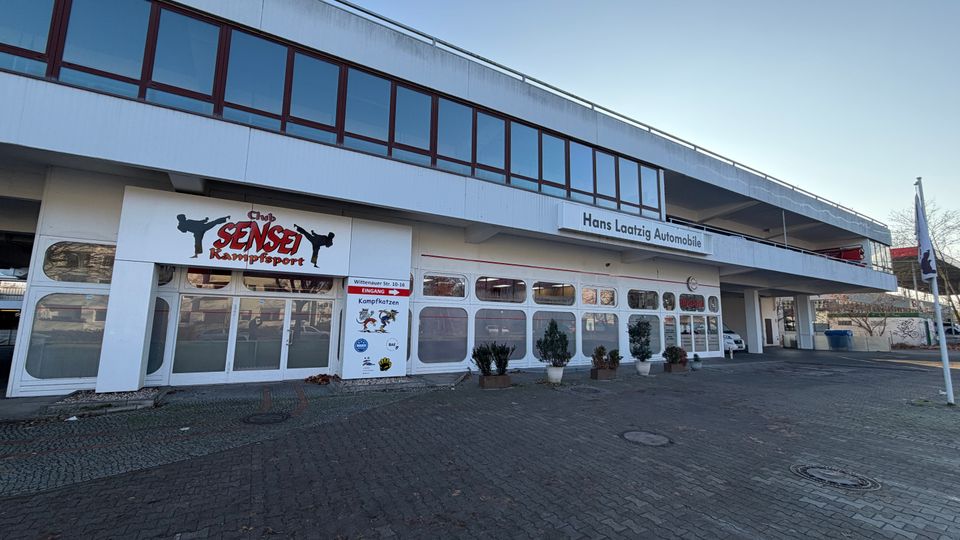 Zweistöckiges Gebäude mit weißer Fassade und großen Fenstern im oberen Bereich, im Erdgeschoss befinden sich ein Kampfsportstudio („Club Sensei Kampfsport“) und ein Autohaus („Hans Laatizg Automobile“). Vor dem Gebäude gibt es einen gepflasterten Vorplatz mit Pflanzkübeln.