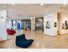 Moderner Empfangsbereich mit Holzfußboden, weißen Wänden und goldgerahmten Glastüren. Im Raum befinden sich Sitzmöbel in Blau und Rot, eine Rezeption, ein blauer Werbeaufsteller mit der Aufschrift „Institut Français“ sowie Wandbilder in Galerieanordnung.