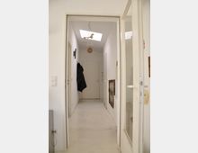 Schmaler, heller Flur in einer Wohnung, mit weißen Wänden, hellem Boden und einer Tür am Ende. An der Decke hängt eine Lampe mit Vogelmotiven, und an den Wänden befinden sich Bilder sowie eine Garderobe mit schwarzen Jacken.